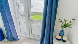 Ein Zimmer mit einer Schiebetür zu einem Garten, einem blauen Vorhang, einer Vase mit Blumen und zwei Kerzen auf einem weißen Tisch.