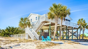 A beachfront house with a wooden deck, a staircase leading to a balcony, and two blue chairs on the sand.