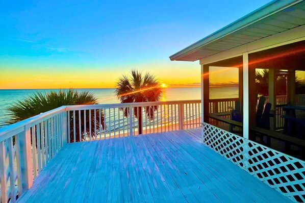 Une terrasse en bois avec une rambarde, donnant sur une plage au coucher du soleil.