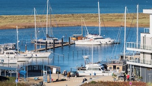 A marina with multiple boats docked, a building with a balcony overlooking the water, and a grassy area in the background. - Vacation apartment Meerblickbude (Wendtorf)