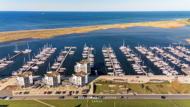 An aerial view of a marina with numerous boats docked, a modern building complex, and a clear view of the ocean. - Vacation apartment Meerblickbude (Wendtorf)