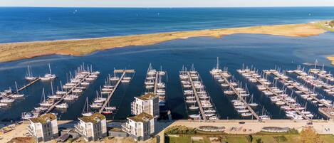 An aerial view of a marina with numerous boats docked, a modern building complex, and a clear view of the ocean.