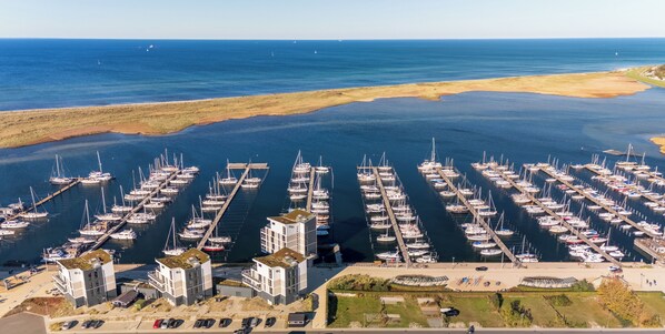An aerial view of a marina with numerous boats docked, a modern building complex, and a clear view of the ocean.