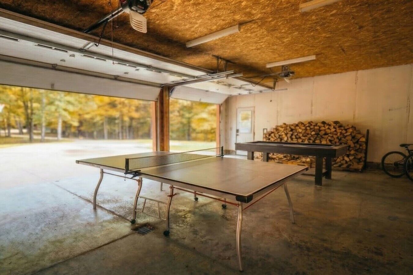 A ping pong table in a garage with a concrete floor, a bicycle, and a stack of firewood.