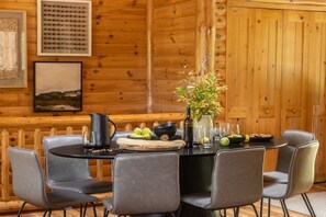 A wooden cabin interior with a round dining table, grey upholstered chairs, a vase with flowers, and framed artwork on the walls.