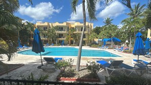 A pool area with lounge chairs and umbrellas, surrounded by palm trees and a building in the background. - Air-conditioned 1 bed 1 full bath 1 half bath apartment.  Hot Water & generator. (Sosúa)