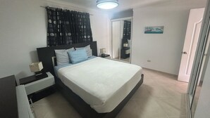 A bedroom with a bed, a nightstand, a ceiling fan, a window with curtains, and a door. - Air-conditioned 1 bed 1 full bath 1 half bath apartment.  Hot Water & generator. (Sosúa)