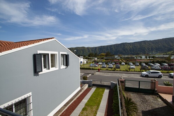A modern building with a grey facade and a red-tiled roof. It features a driveway leading to a parking area with cars and a view of a mountainous landscape in the background.