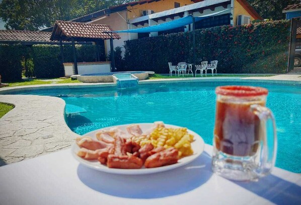 A poolside table with a plate of food and a glass of beverage, surrounded by a pool and outdoor seating. - Hotel Casa Kolping (Tuxtla Gutierrez)