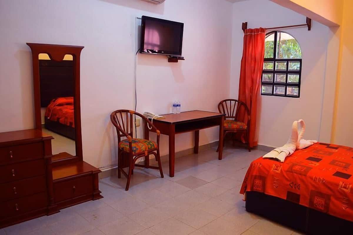 Quarto com cama, escrivaninha, duas cadeiras, televisão e janela com cortinas.