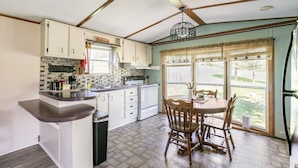 A compact kitchen with white cabinets, a wooden dining table, and a large window offering a view of the outdoors. - Dorothy’s Retreat (Campbellsville)