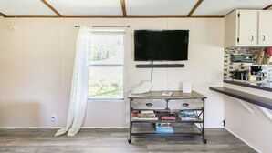 A compact kitchen with a flat-screen TV, a small dining table, and a window with a curtain. - Dorothy’s Retreat (Campbellsville)