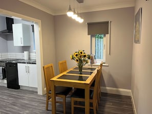 A dining area with a table, chairs, a vase with sunflowers, a window with blinds, and a kitchen in the background.