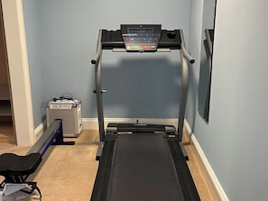 A treadmill in a room with light blue walls, a carpeted floor, and a small piece of exercise equipment in the corner.
