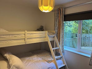 A bunk bed with a teddy bear on the top bunk, a window with a view of a wooden fence, and a patterned curtain.