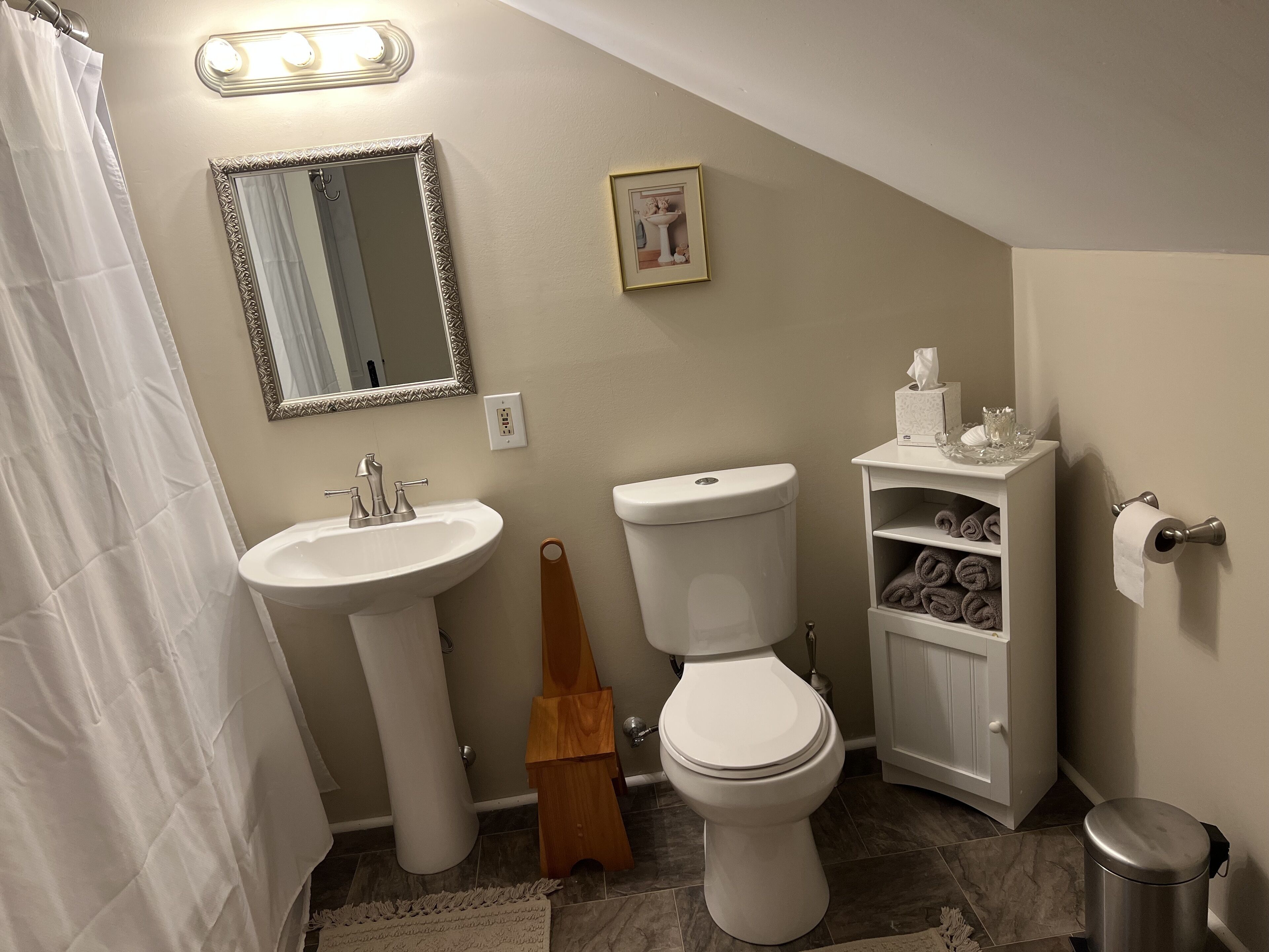 Et badeværelse med håndvask på piedestal, toilet og et lille skab med håndklæder.