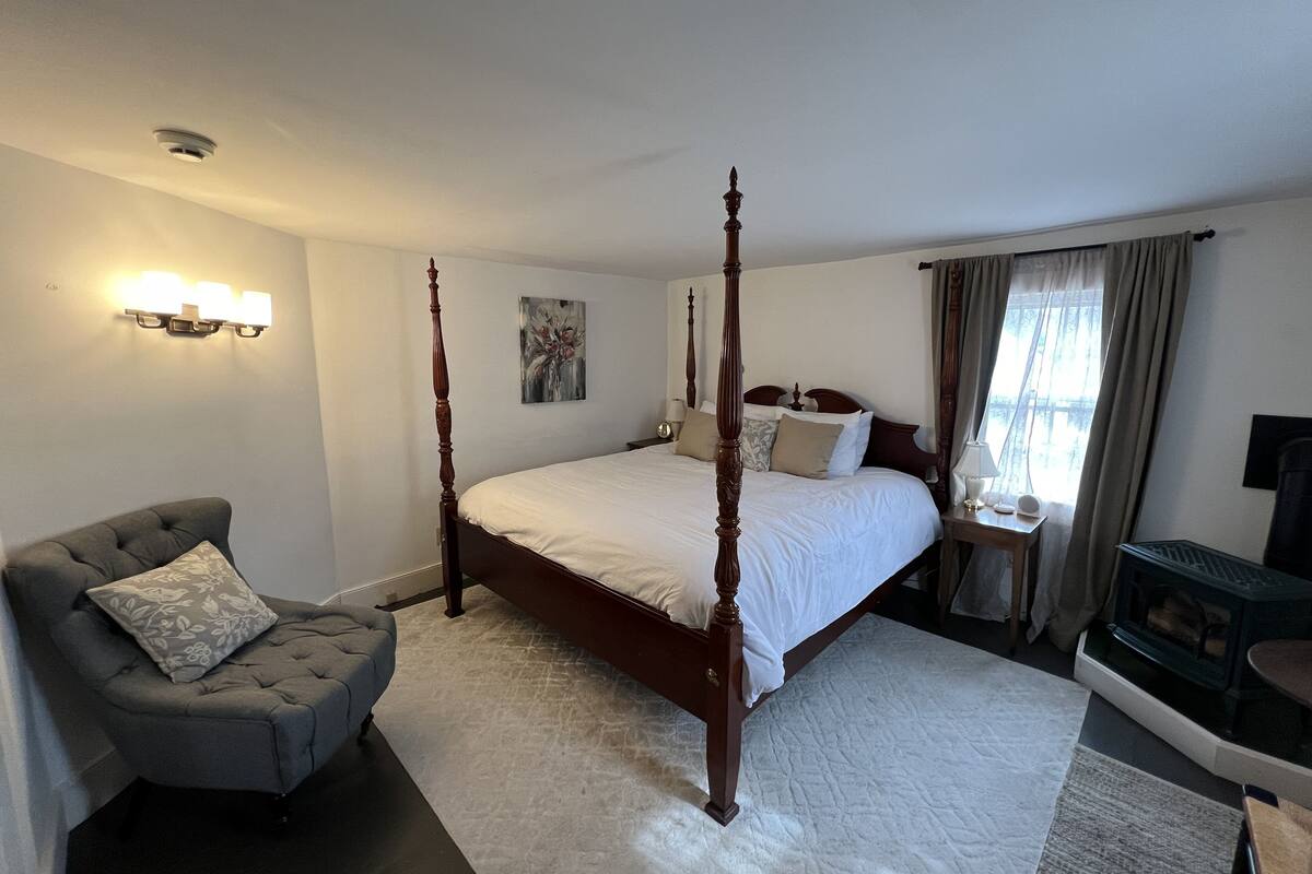 A four-poster bed with white linens, a grey armchair with a floral pillow, a bedside table with a lamp, and a painting on the wall.