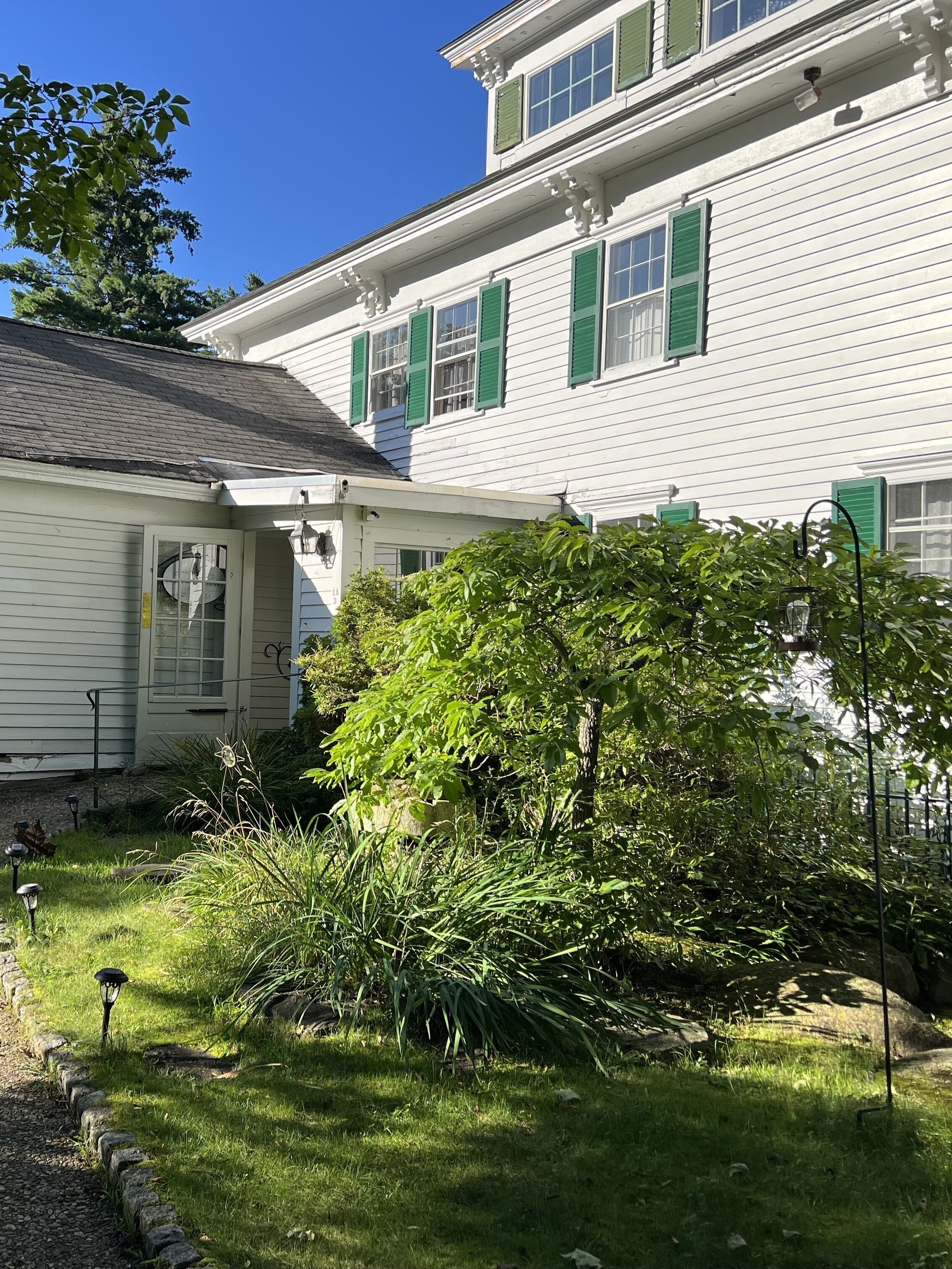 A white building with green shutters, a covered porch, and a garden with various plants and a pathway.