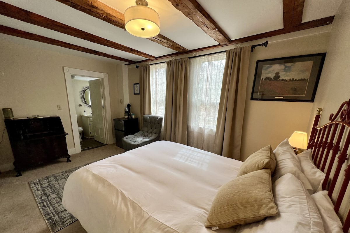 A bedroom with a large bed, a chair, a desk, and a window with curtains.