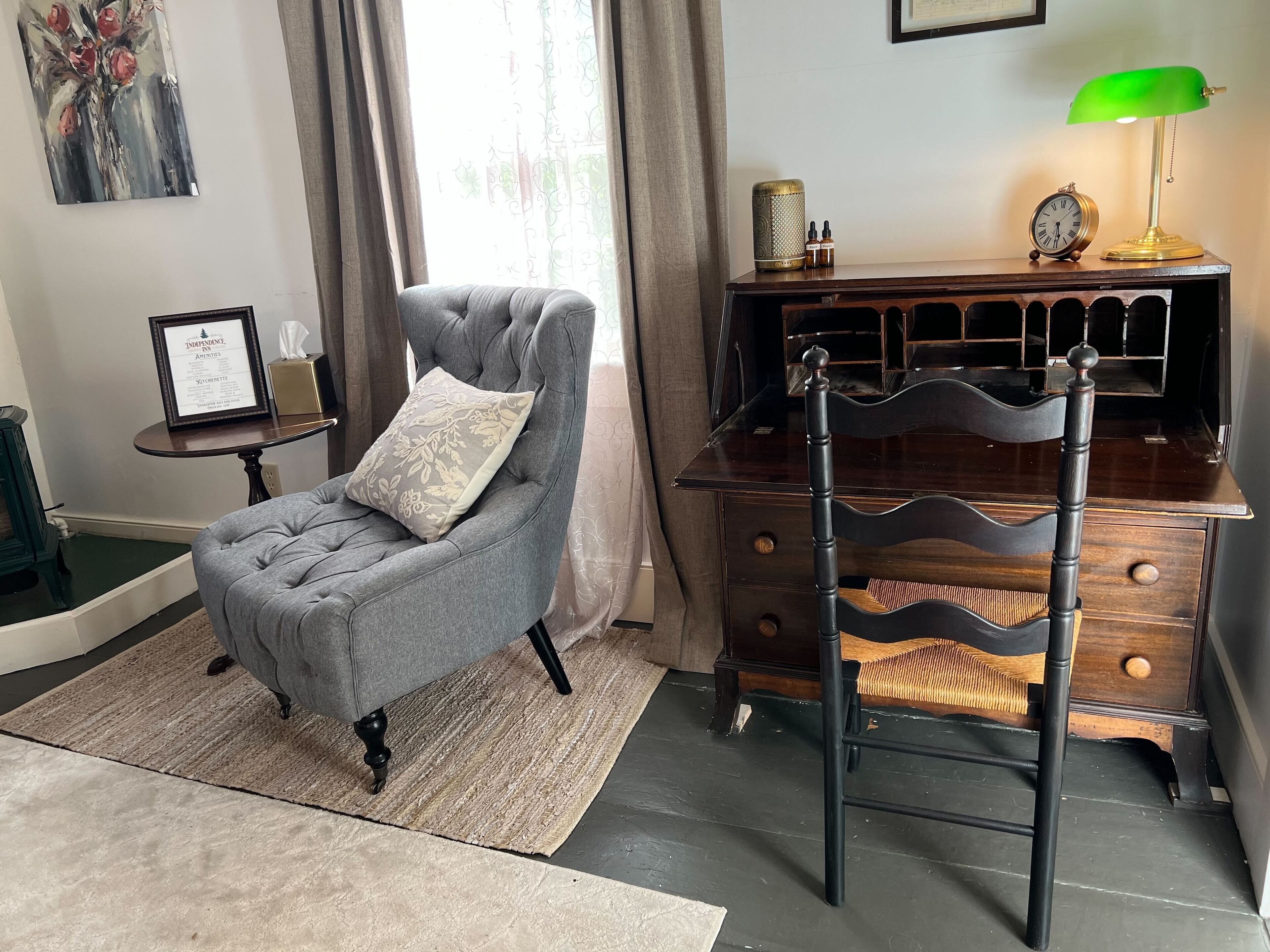 A room with a grey armchair, a wooden dresser, a green desk lamp, and a framed certificate on a small table.
