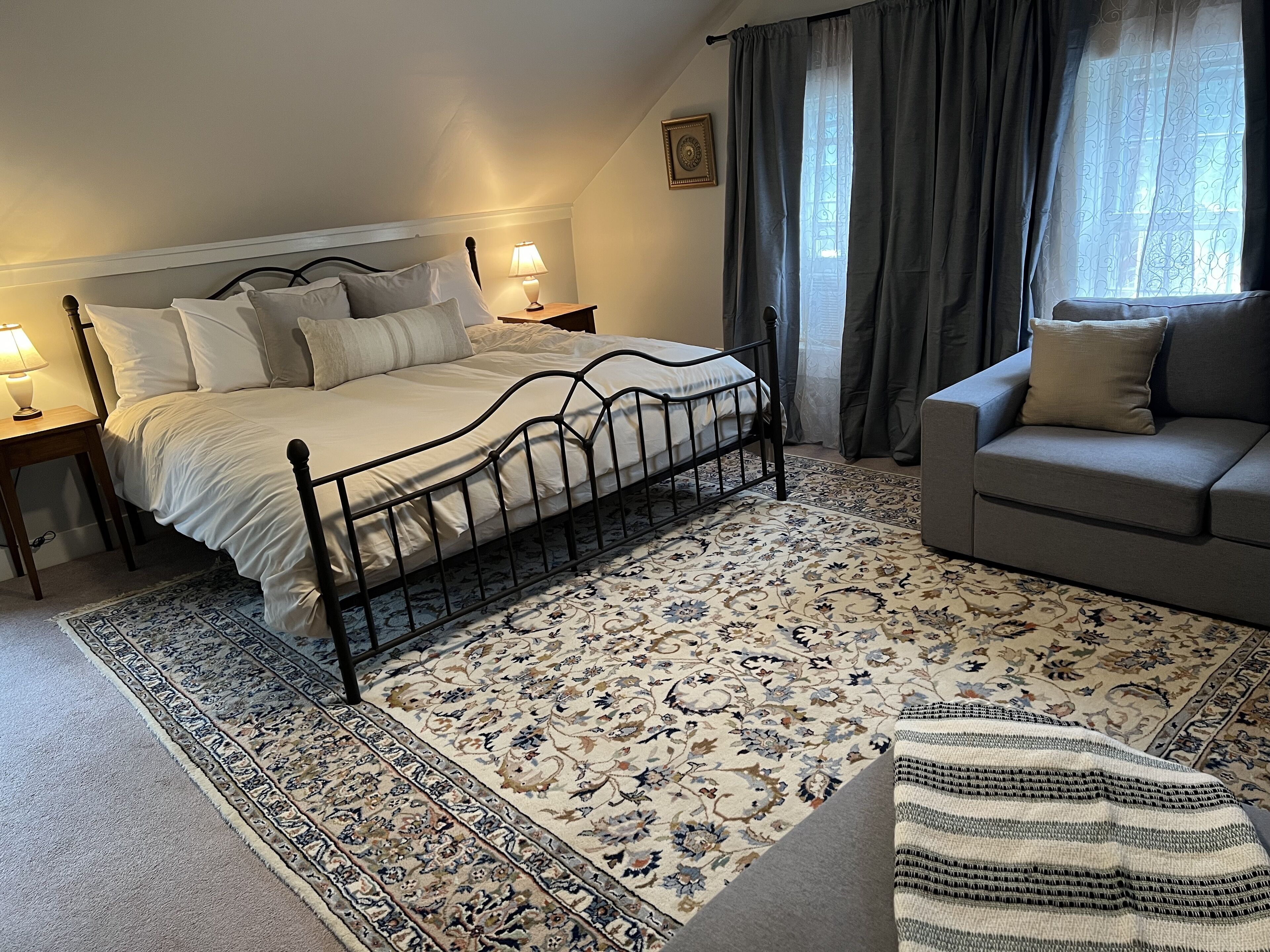 Un dormitorio con cama, mesitas de noche, un sofá, alfombra estampada y una ventana con cortinas.