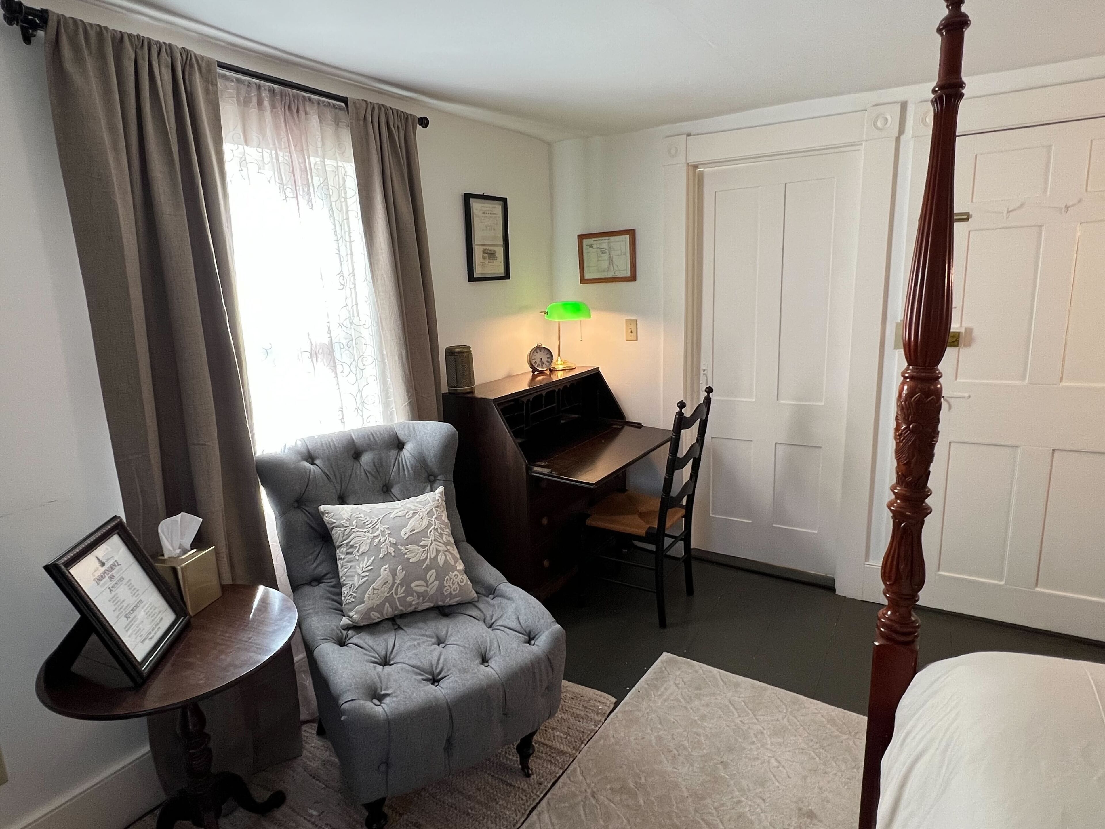 A bedroom with a four-poster bed, a grey armchair with a cushion, a small table with a lamp, and a desk with a chair.