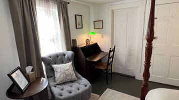 A bedroom with a four-poster bed, a grey armchair with a cushion, a small table with a lamp, and a desk with a chair.