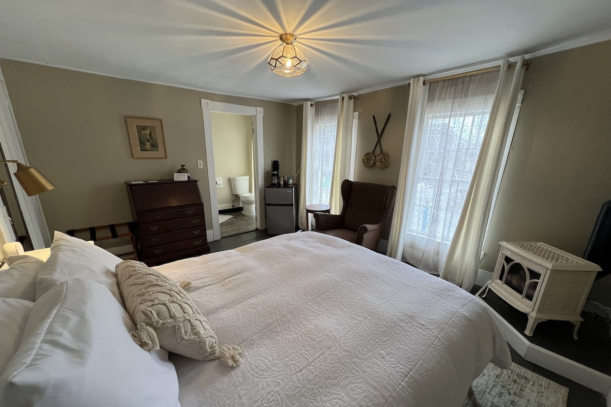 A hotel room with a large bed, a dresser, a chair, a small table, a heater, and a window with curtains.