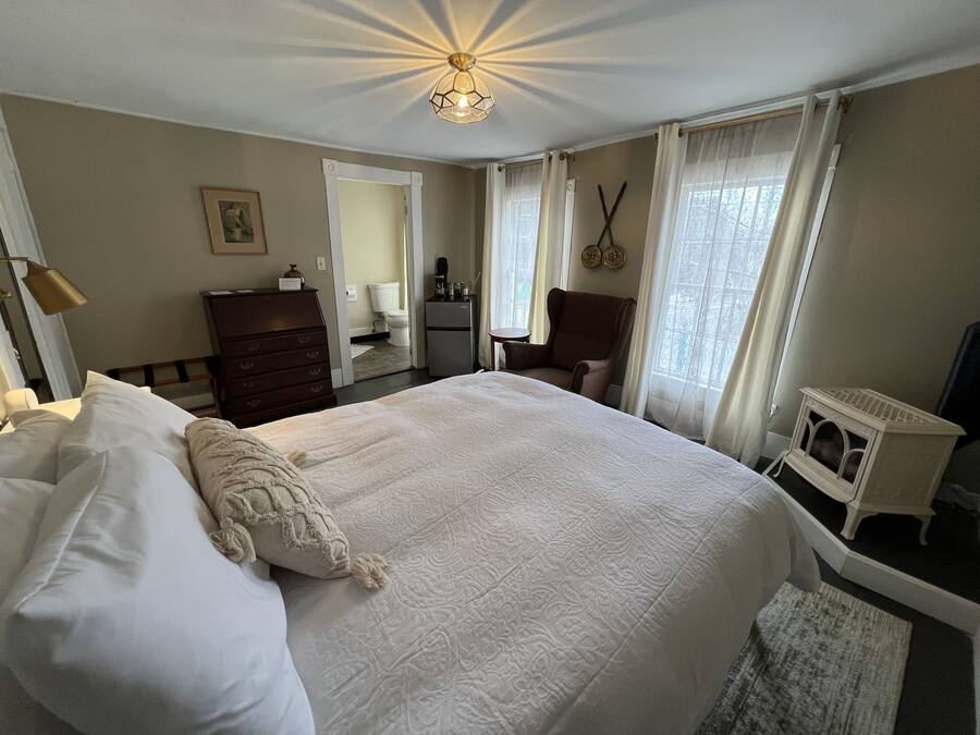 Une chambre d’hôtel avec un grand lit, une commode, une chaise, une petite table, un radiateur et une fenêtre avec des rideaux.