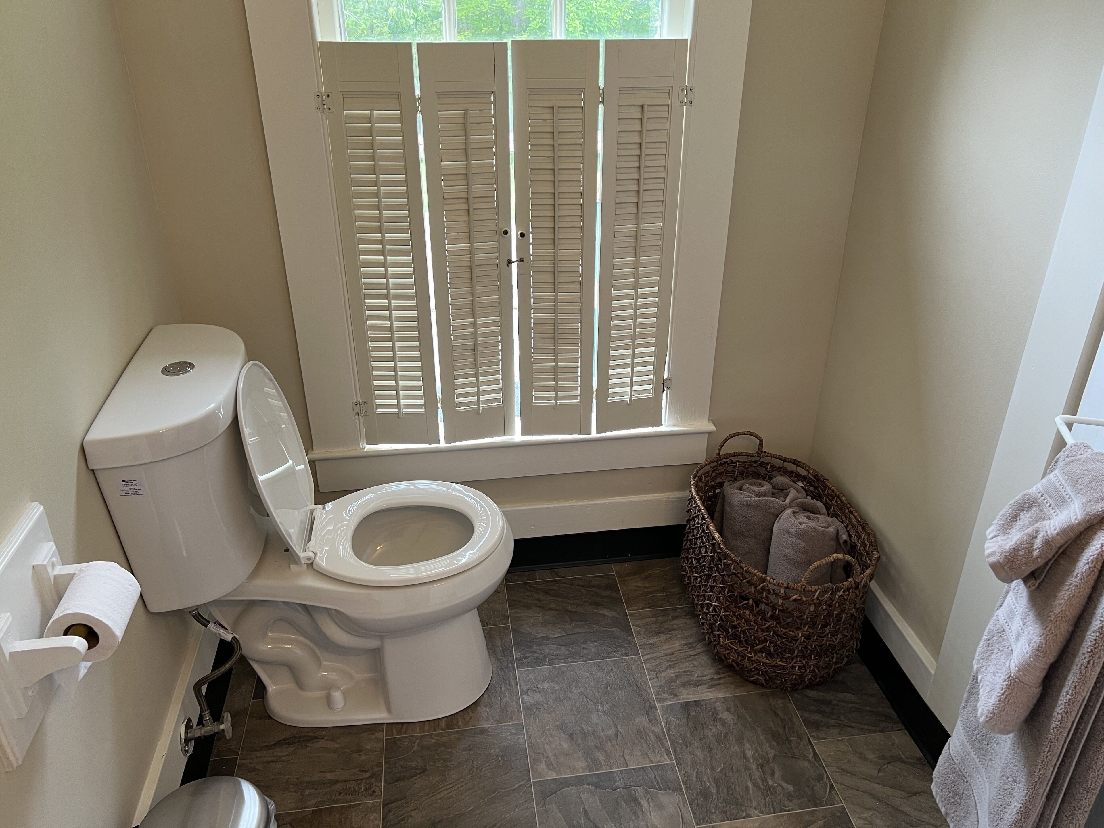 A bathroom with a toilet, a window with shutters, a wicker basket, and a towel rack.