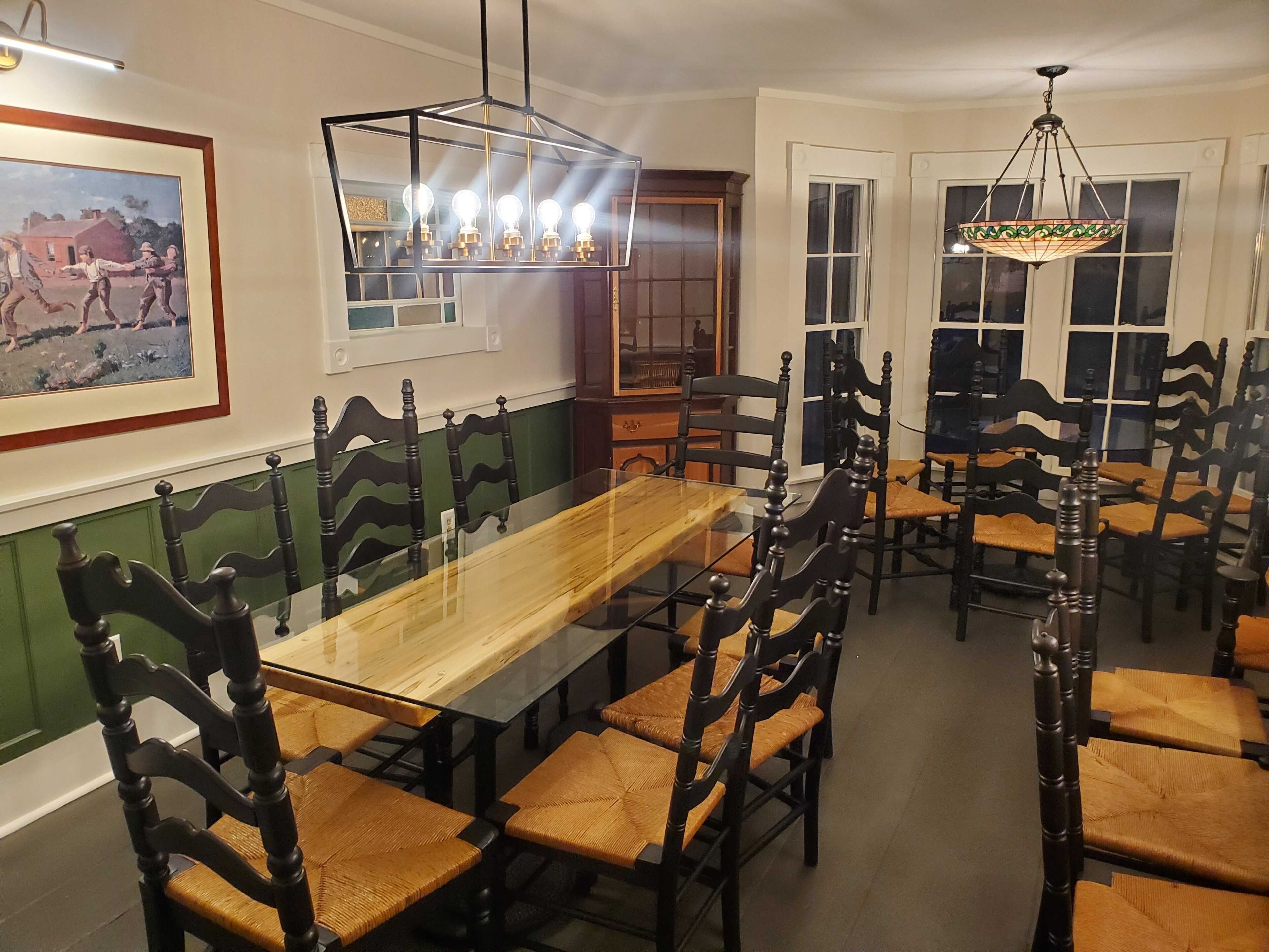 A dining area with a long wooden table surrounded by dark wooden chairs with woven seats. There is a framed picture on the wall and a hanging light fixture above the table.