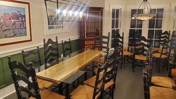 A dining area with a long wooden table surrounded by dark wooden chairs with woven seats. There is a framed picture on the wall and a hanging light fixture above the table.
