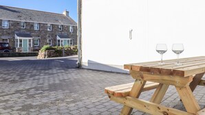 Une table de pique-nique en bois avec deux verres à vin posés sur une surface pavée, un bâtiment en pierre avec des fenêtres et une porte, et un ciel bleu dégagé.