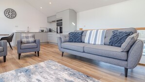 Un salon moderne avec un canapé gris, deux fauteuils, une table basse et une cuisine en arrière-plan.