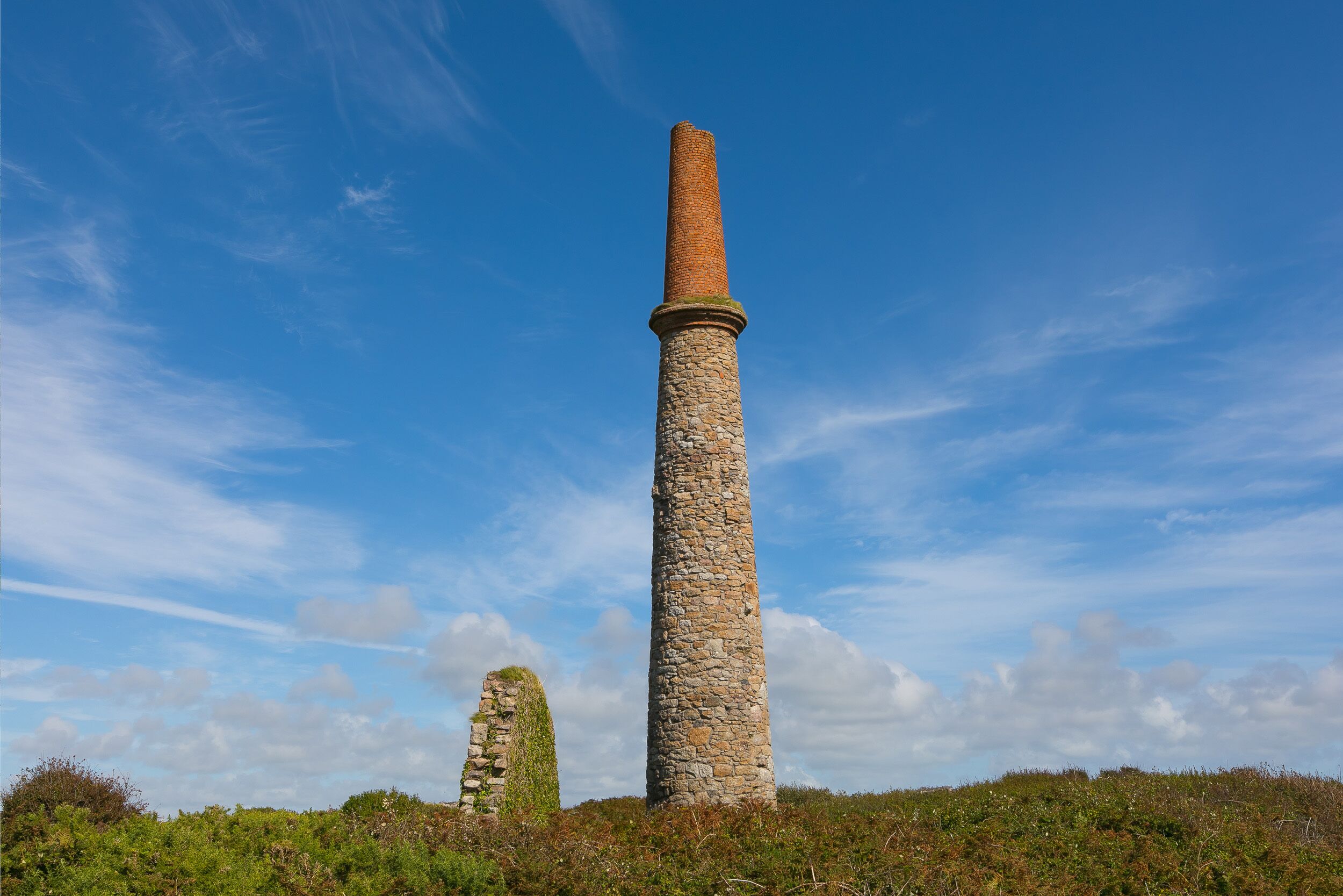 A tall stone structure with a brick chimney, surrounded by greenery and a clear blue sky.