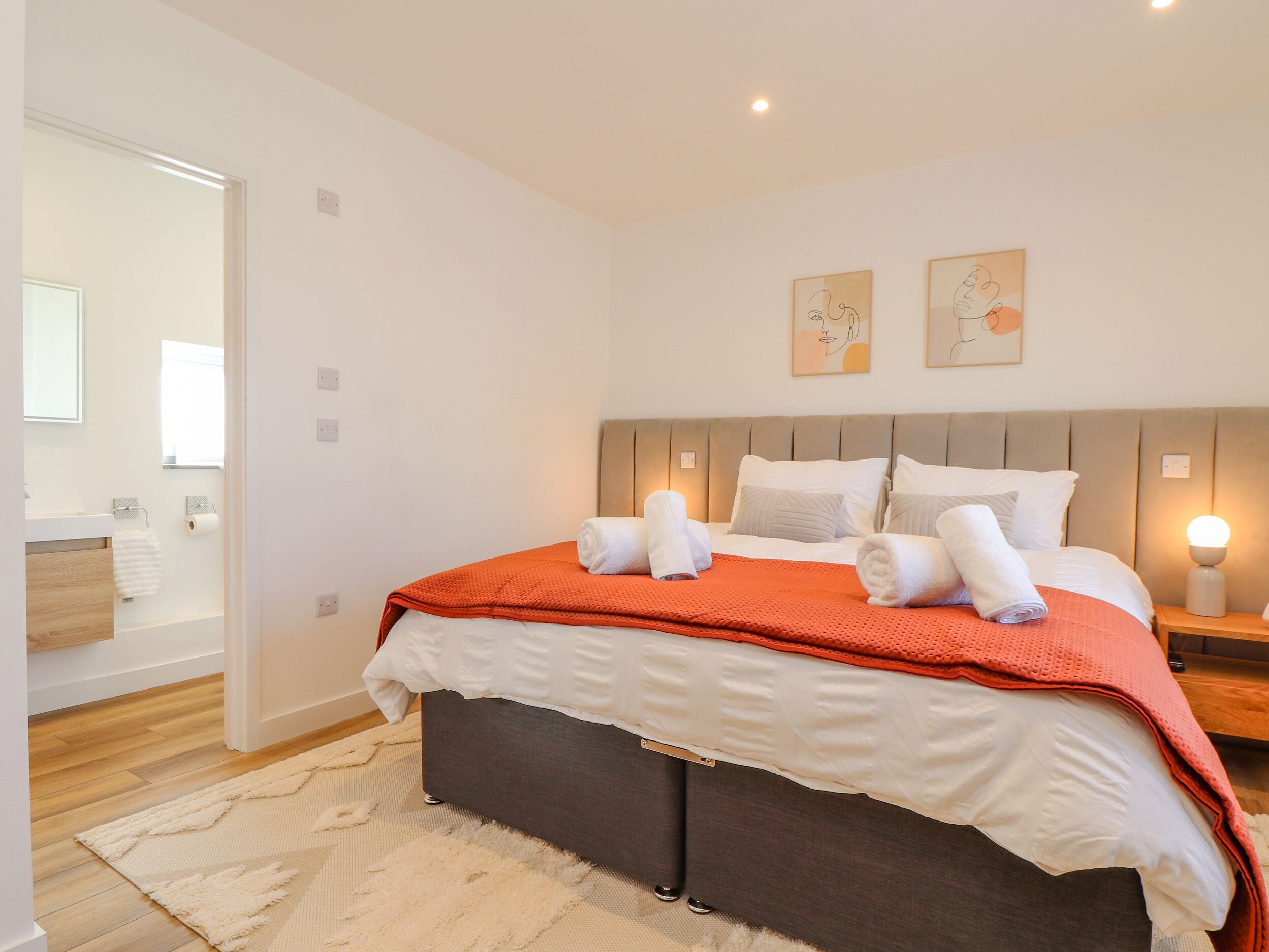 Un dormitorio con cama, mesita de noche, lámpara de noche, cabecera y baño visible a través de una puerta abierta.