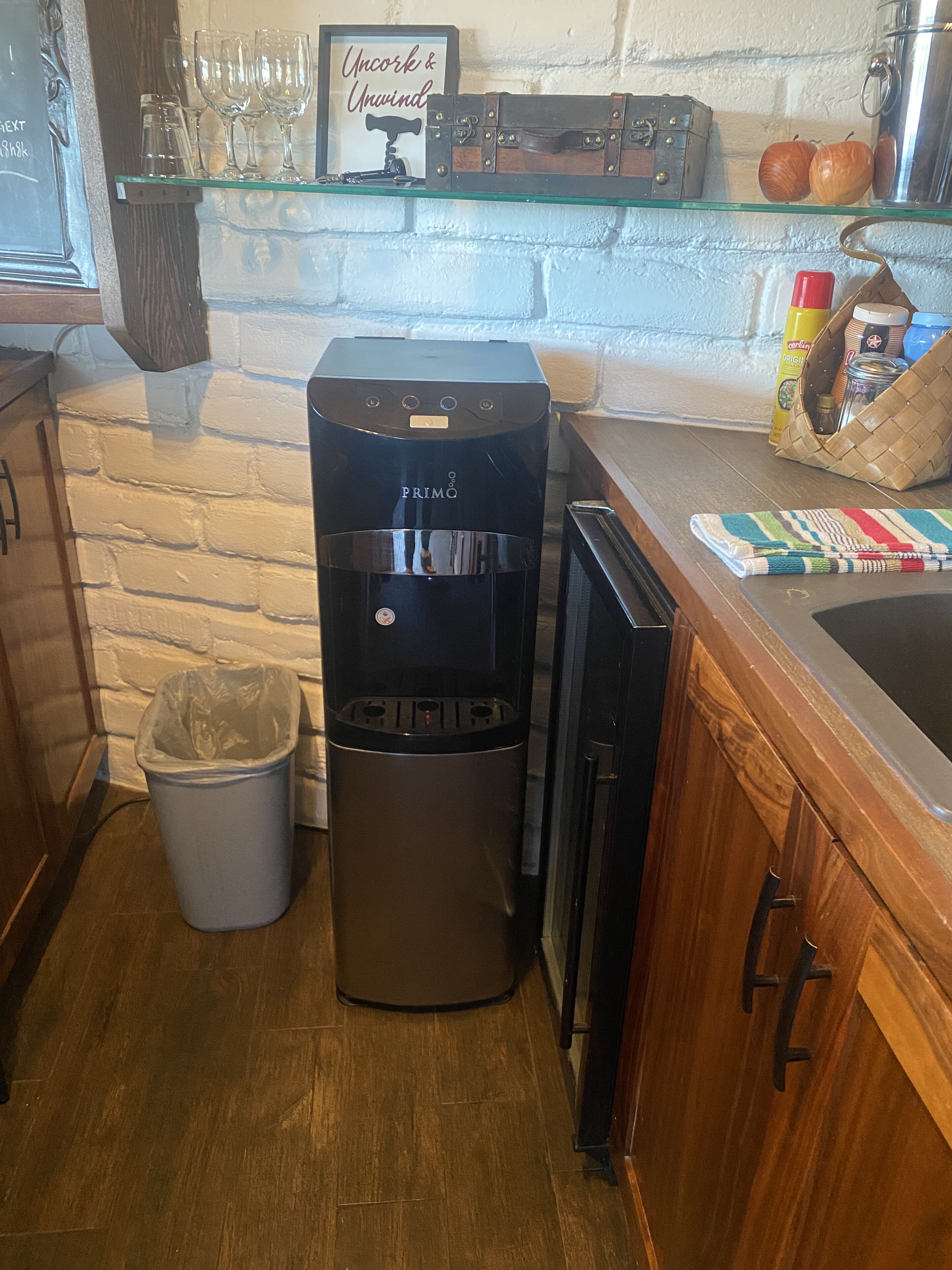 Une petite cuisine équipée d’une machine à café, d’une poubelle et d’une étagère où sont rangés des verres et un cadre avec une inscription.