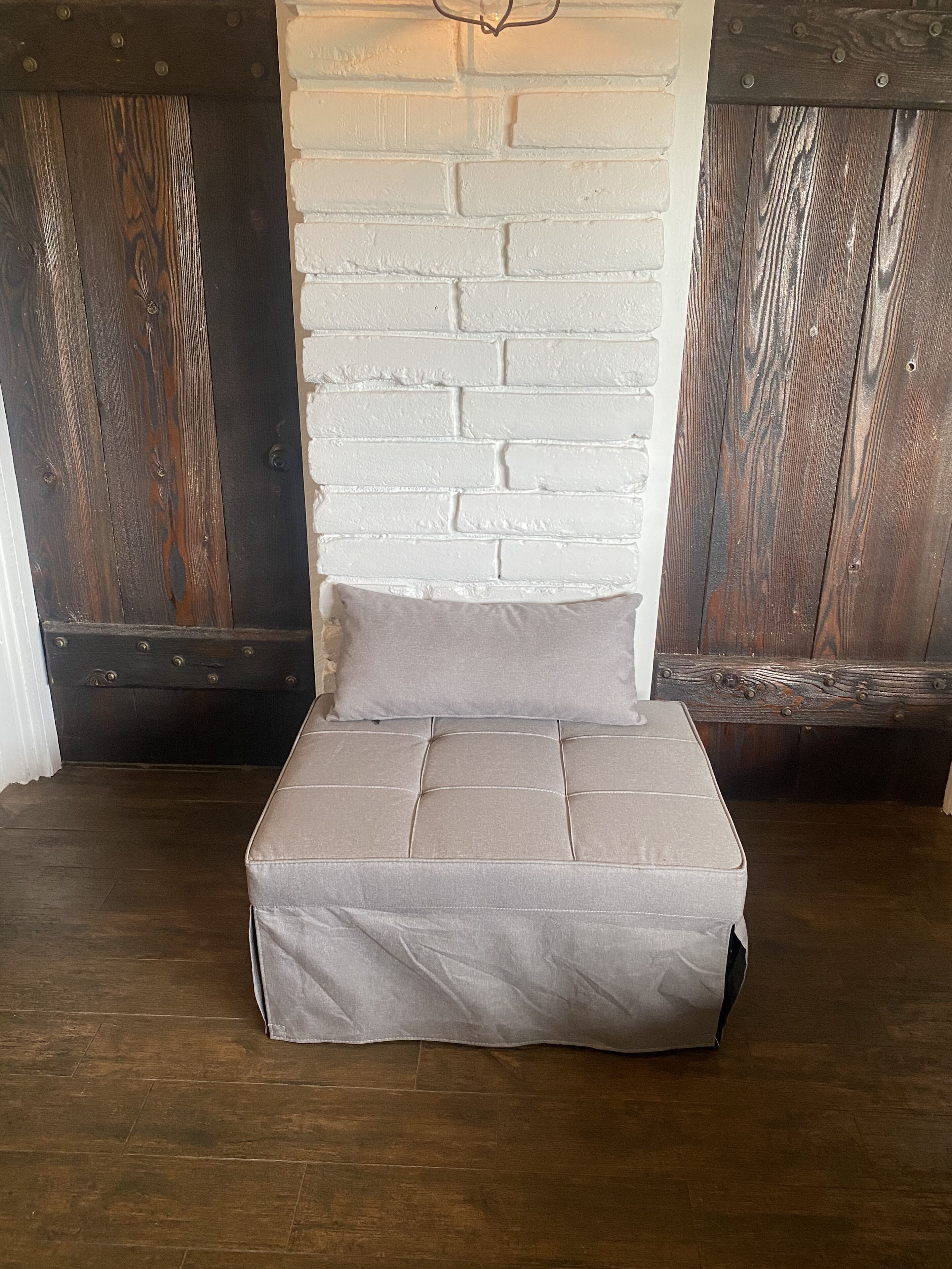 A small, square bed with a grey mattress and pillow, positioned against a white brick wall with wooden paneling on either side.