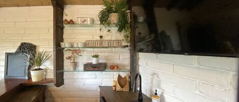 Uma cozinha com paredes de tijolos brancos, teto de madeira e prateleiras com diversos objetos, incluindo plantas, uma placa de café e utensílios de cozinha.