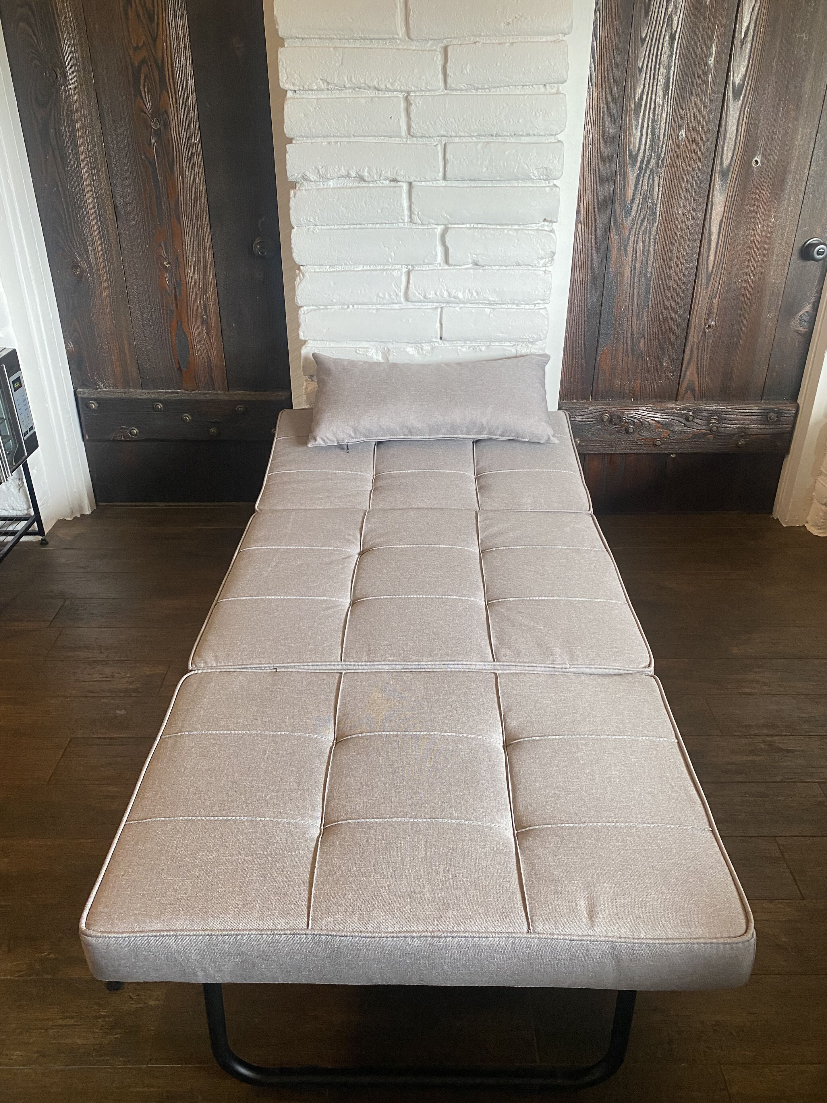 Un letto singolo con materasso grigio e struttura in metallo nero in una stanza con pavimento in legno e parete in mattoni bianchi.