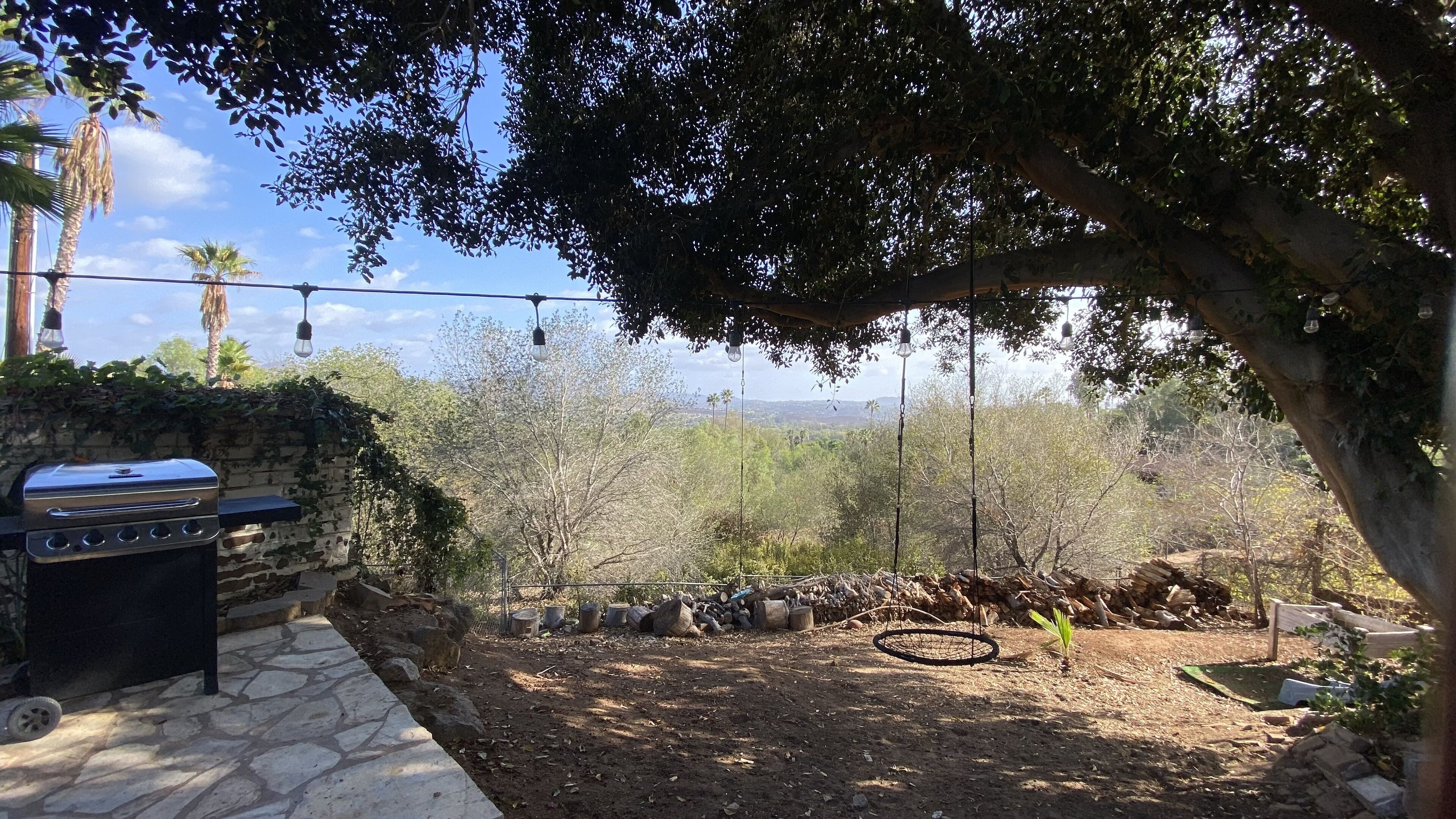 A backyard with a barbecue grill, a stone pathway, and a view of trees and hills.