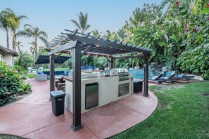 An outdoor kitchen area with a pergola, seating, and a grill. - Luxury Retreat-Pool/HotTub/Game Room/Putting Green (Escondido)