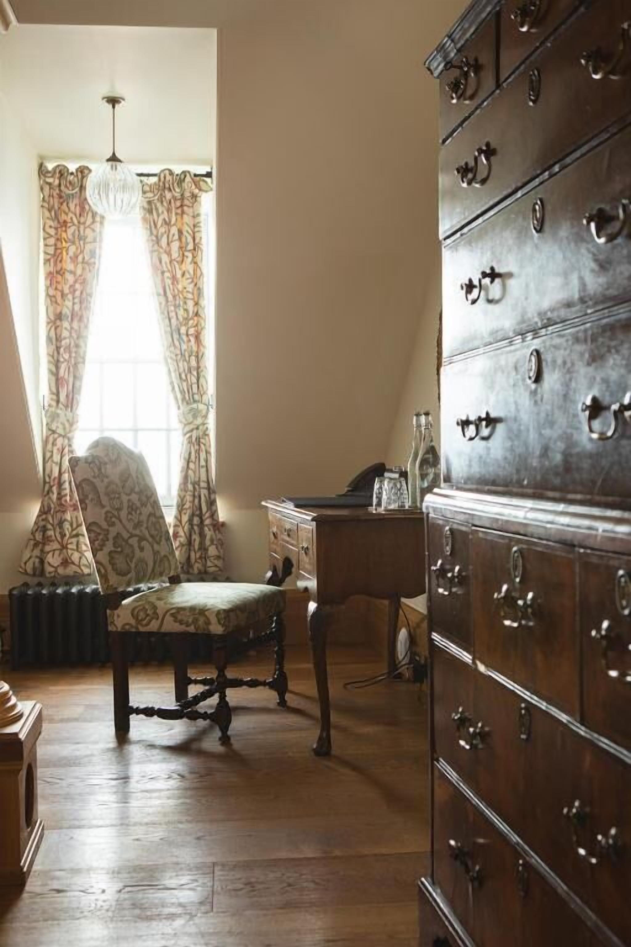 Une pièce avec un plancher en bois, une chaise de style vintage, une grande commode en bois dotée de nombreux tiroirs, une petite table et une fenêtre ornée de rideaux à motifs floraux.