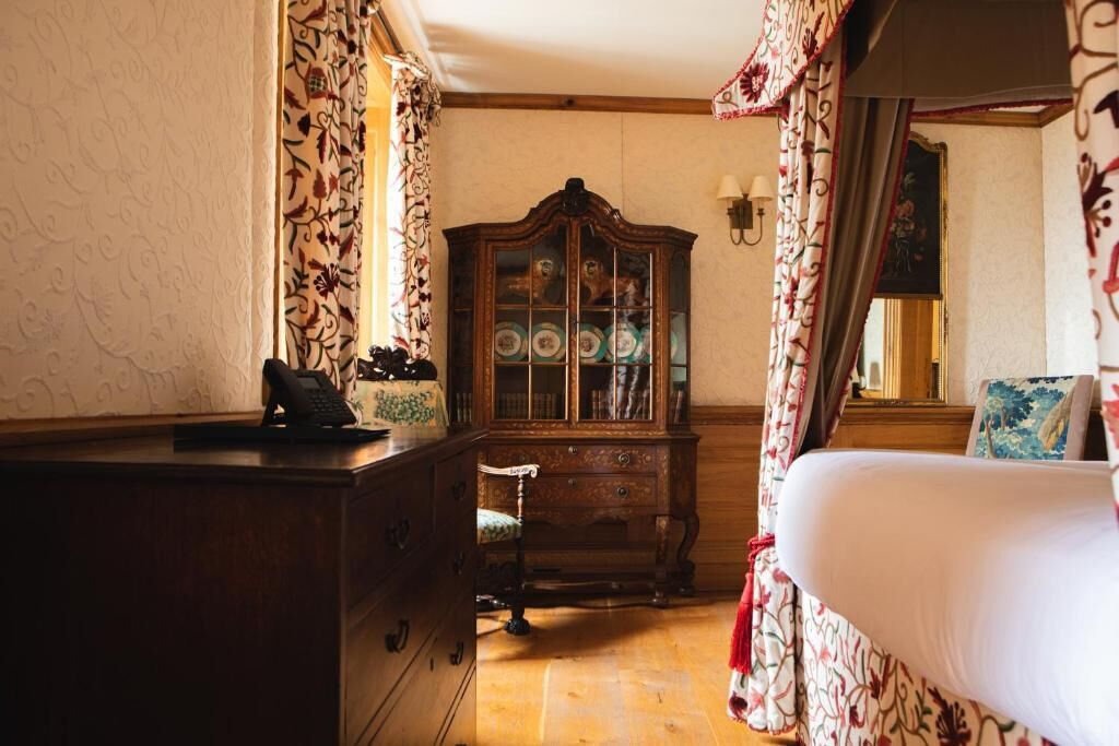 A traditional room with a wooden dresser, a cabinet with glass doors, a bed with floral curtains, and a chair.