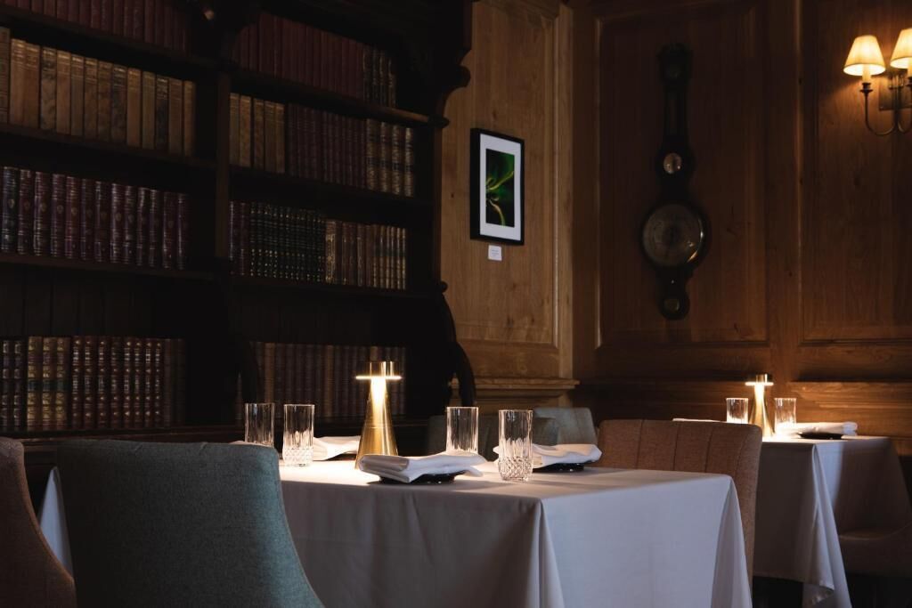 Een kamer in gedimd licht met een grote houten boekenkast vol boeken, een hangklok en een ingelijste foto. Tafels zijn gedekt met kaarsen en glazen.
