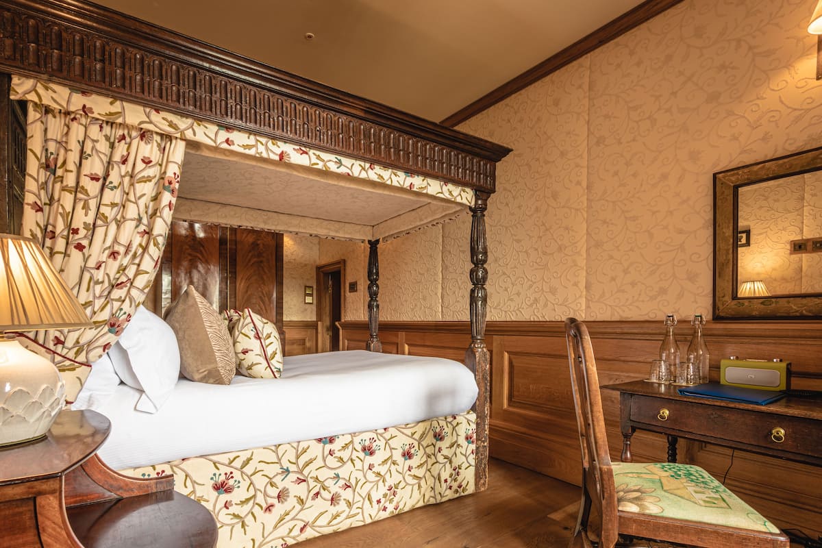 A four-poster bed with a floral-patterned upholstered headboard, a wooden nightstand with a lamp, and a chair with a floral cushion.