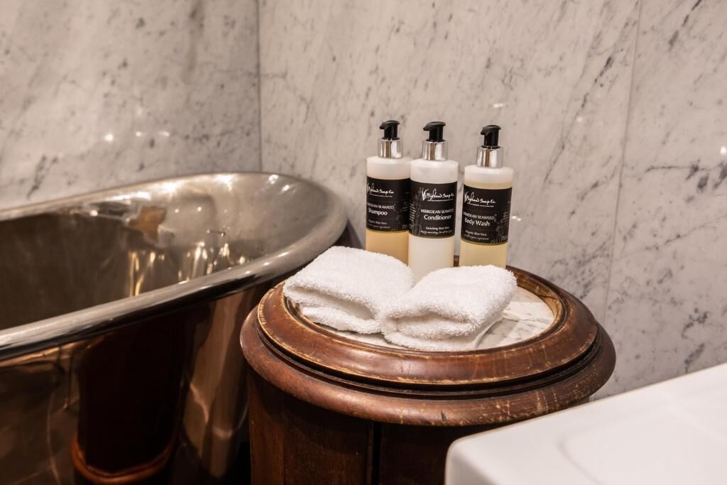 A bathroom with a marble wall, a bathtub, and three bottles of body wash and conditioner on a wooden stool.