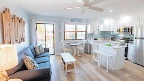 Eine moderne Küche mit weißen Schränken, einer zentralen Kochinsel und Edelstahlgeräten. Es gibt einen Essbereich mit Tisch und Stühlen sowie ein Wohnzimmer mit Sofa und Lampe.