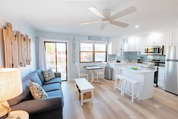 A modern kitchen with white cabinets, a central island, and stainless steel appliances. There is a dining area with a table and chairs, and a living room with a sofa and a lamp.
