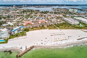 Vue aérienne d’une ville côtière avec une grande plage de sable, un port de plaisance et divers bâtiments, notamment des hôtels et des zones résidentielles.
